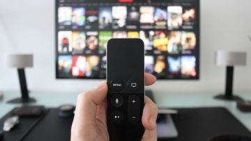 Imagen de una persona que sostiene el control de un televisor con la mano, mientras está frente a una pantalla con opciones de series y películas.