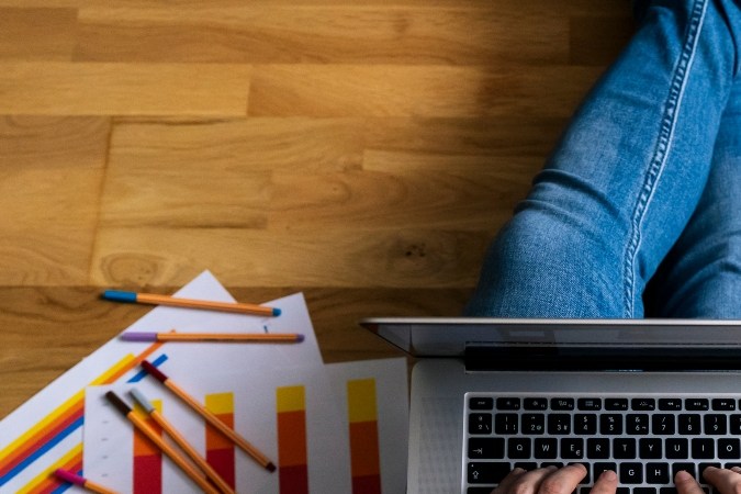 Imagen de una persona que está sentada en el suelo con una computadora en las piernas y varias hojas alrededor.