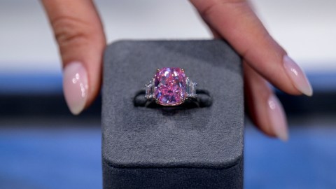 Imagen de un diamante de color rosa que está montado en un anillo de color plata, sujeto a una caja de color gris, que es sostenida por tres dedos.
