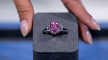 Imagen de un diamante de color rosa que está montado en un anillo de color plata, sujeto a una caja de color gris, que es sostenida por tres dedos.