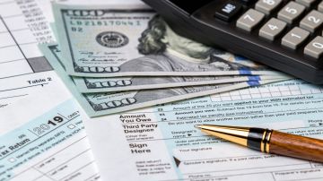 Imagen de tres billetes debajo de una calculadora, sobre varios formularios de impuestos y una pluma.
