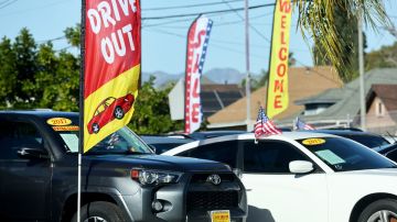 Imagen de dos autos, una camioneta de color gris y un auto de color blanco, que están estacionados entre banderas de color rojo y amarillo, en un sitio de venta de autos usados.