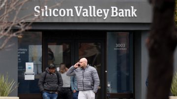Imagen de la entrada del banco Silicon Valley Bank, en color gris, y de una persona con una chamarra gris que habla por teléfono.