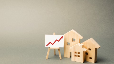 Imagen de varias casas en miniatura de color madera que están junta a una pizarra con una flecha de color rojo ascendente.