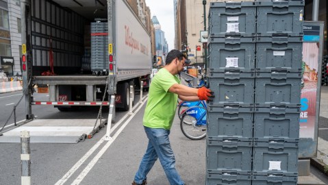 Imagen de una persona que empuja una pila de cajas de plástico y en el fondo un tráiler estacionado en la calle.