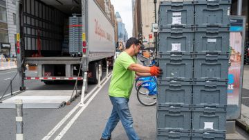 Imagen de una persona que empuja una pila de cajas de plástico y en el fondo un tráiler estacionado en la calle.