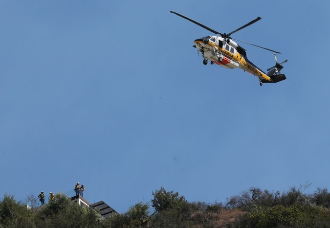 LOS ANGELES, CALIFORNIA - OCTOBER 21: A Los Angeles County Fire Department helicopter flies above firefighters positioned on a rooftop during the Palisades Fire, in the Pacific Palisades neighborhood, on October 21, 2019 in Los Angeles, California. The fire scorched at least 40 acres and threatened hillside homes. (Photo by Mario Tama/Getty Images)
