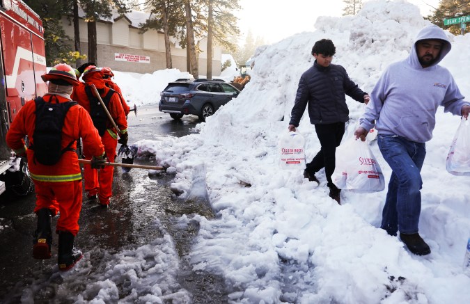 CRESTLINE, CALIFORNIA - MARCH 03: A crew of inmate firefighters (L) walk back to their vehicle after shoveling and clearing snow, while people carry bags of food (R), after a series of winter storms in the San Bernardino Mountains in Southern California on March 3, 2023 in Crestline, California. Some residents have been stranded in Crestline more than a week due to the snowfall while the local grocery store was severely damaged when its roof collapsed under the weight of the snow. California Governor Gavin Newsom has declared a state of emergency due to winter storms for 13 counties including San Bernardino County and the California National Guard is assisting relief efforts in the mountains. (Photo by Mario Tama/Getty Images)