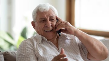 Un adulto mayor se sienta en el sofá de casa y se ríe hablando por un teléfono celular moderno.