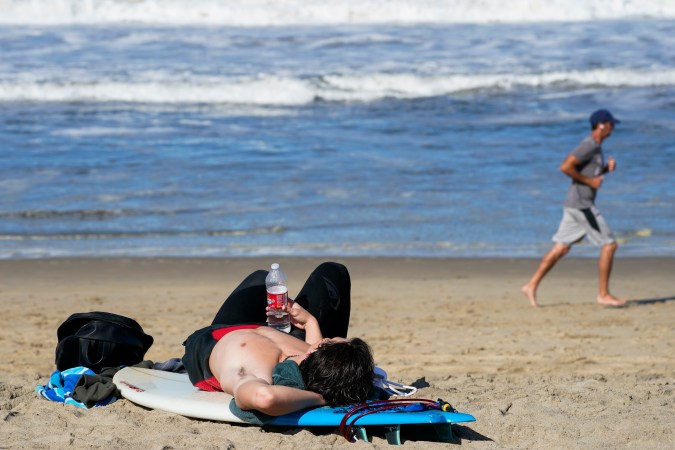 A surfer rests on his board as a jogger passes on Wednesday, May 13, 2020, in Manhattan Beach, Calif. Los Angeles County reopened its beaches Wednesday in the latest cautious easing of coronavirus restrictions that have closed most California public spaces and businesses for nearly two months. (AP Photo/Ashley Landis)
