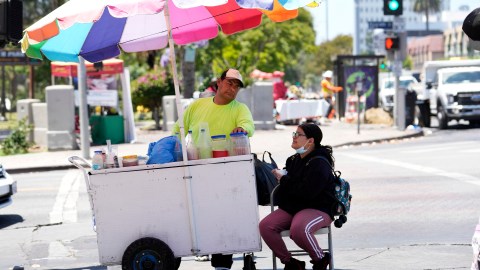 A street vendor selling snow cones tends to a customer at a street corner near MacArthur Park, Tuesday, July 11, 2023, in Los Angeles. (AP Photo/Marcio Jose Sanchez)