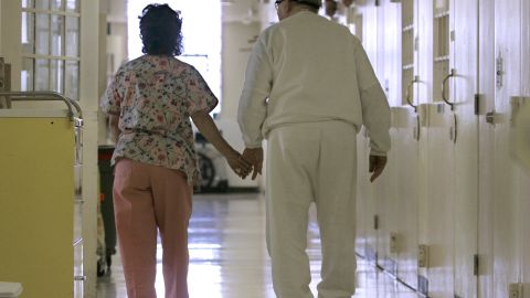 FILE - In this Wednesday, April 9, 2008 file photo, Debbie Coluter, a certified nursing assistant, holds the hand of an elderly inmate with Alzheimer's disease, as she helps him to his cell at the California Medical Facility in Vacaville, Calif. (AP Photo/Rich Pedroncelli)