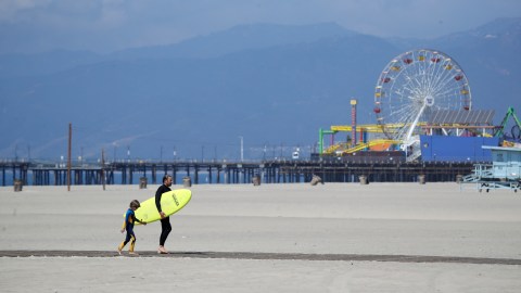 A surfer with a child walks on the mostly empty Santa Monica beach Sunday, March 29, 2020, in Los Angeles. With cases of coronavirus surging and the death toll increasing, lawmakers are pleading with cooped-up Californians to spend a second weekend at home to slow the spread of the infections. (AP Photo/Marcio Jose Sanchez)