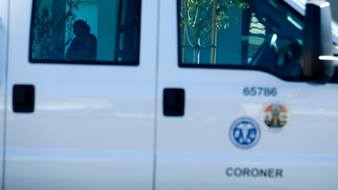 A neighbor, back left, stands behind a Los Angeles County Coroner vehicle parked near a home where five bodies were found in the city of Lancaster in the high desert Antelope Valley north of Los Angeles, Monday, Nov. 29, 2021. A Los Angeles County Sheriff's Department statement says deputies found a woman, a girl and three boys with gunshot wounds and paramedics pronounced them dead at the scene. The department says the children's father showed up at the Lancaster sheriff's station and was arrested on suspicion of five murders after being interviewed by detectives. (AP Photo/Ringo H.W. Chiu)