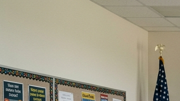 Aula pública en Texas con pizarrón, bandera de EE.UU. y un cartel religioso al fondo, sin que domine toda la imagen.