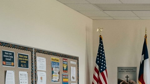 Aula pública en Texas con pizarrón, bandera de EE.UU. y un cartel religioso al fondo, sin que domine toda la imagen.