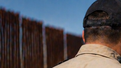 Oficial de seguridad deteniendo a un hombre cerca de la frontera en Texas junto a una patrulla y muro fronterizo.