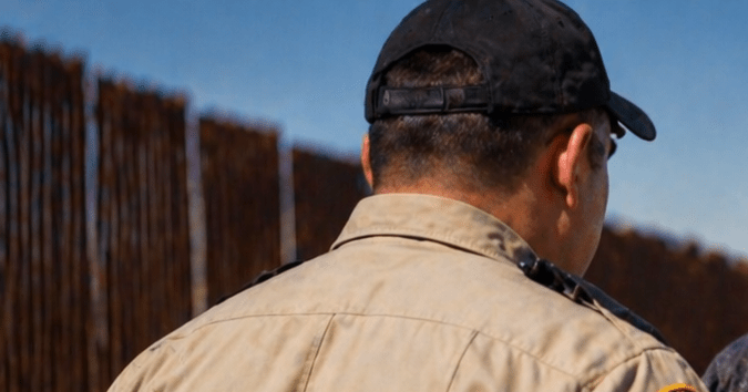 Oficial de seguridad deteniendo a un hombre cerca de la frontera en Texas junto a una patrulla y muro fronterizo.