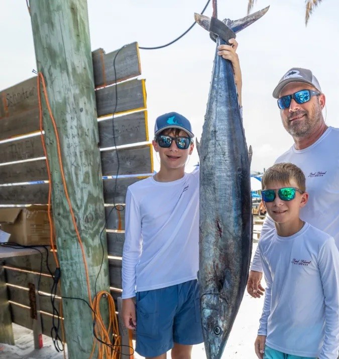 Un padre y sus dos hijos posan junto a un pez peto colgado en la estación de pesaje del Flora-Bama Fishing Rodeo, con la playa de Perdido Key al fondo.