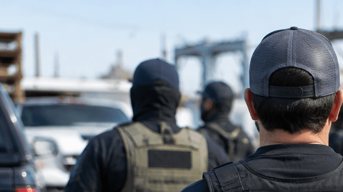 Agente de ICE durante un operativo en un área industrial con trabajadores reunidos al fondo