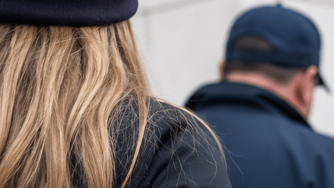 Mujer vista de espaldas mientras es escoltada por agentes en una escena exterior.