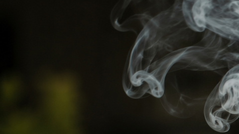 Primer plano de un hombre joven fumando un porro, con humo visible saliendo del cigarrillo y fondo desenfocado.